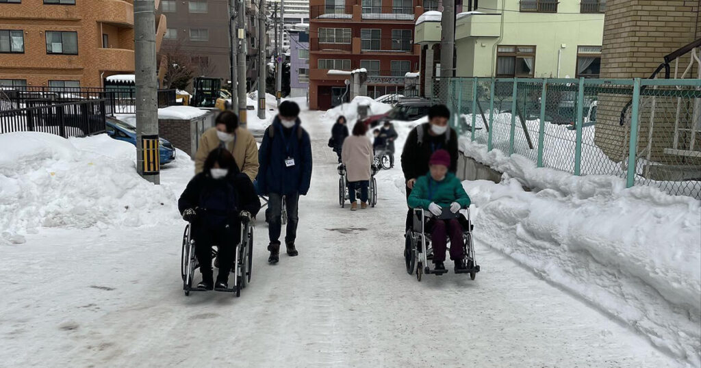 雪の坂道で車椅子を押す受講生達